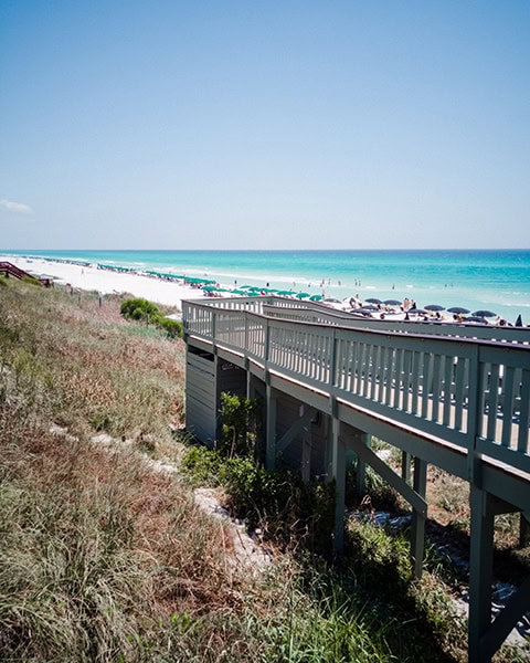 Ein grauer Holzsteg führt über bewachsene Dünen hinweg direkt zum feinen Sandstrand am Henderson Beach State Park in Destin, Florida, an dem sich zahlreiche grüne und dunkle Sonnenschirme entlang des türkisfarbenen Meeres unter einem klaren blauen Himmel aneinanderreihen.
