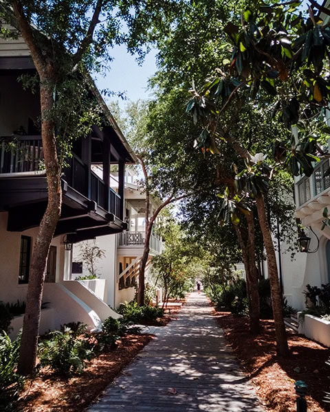 Ein schattiger Fußgängerweg aus Holzbohlen führt flankiert von hohen, grünen Laubbäumen durch die idyllische Wohn- und Ferienhausanlage von Rosemary Beach in Florida, während links und rechts die typischen hellen Fassaden mit dunklen Holzbalkonen im kühlen Schatten liegen.