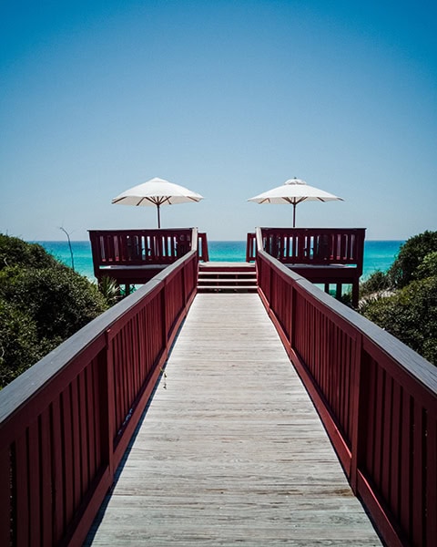 Ein langer Holzsteg mit rötlich-braunem Geländer führt durch dichte Dünenbepflanzung direkt zu einer geteilten Aussichtsplattform mit zwei weißen Sonnenschirmen im Gulf State Park in Gulf Shores, von der aus sich ein weiter Blick auf das türkisfarbene Meer des Golfs von Mexiko unter einem klaren blauen Himmel bietet.