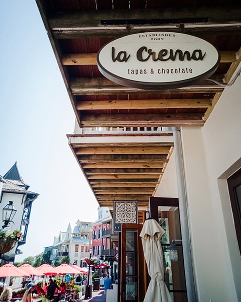 Das ovale Werbeschild des Restaurants La Crema Tapas & Chocolate hängt unter einem hölzernen Balkonüberhang an einer hellen Fassade, während im Hintergrund die von europäischen Baustilen inspirierte Fußgängerzone von Rosemary Beach in Florida mit roten Sonnenschirmen und Urlaubern zu sehen ist.