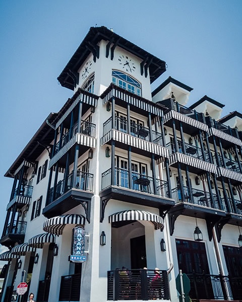 Das markante, mehrstöckige Gebäude des luxuriösen The Pearl Hotel in Rosemary Beach, Florida, zeigt eine weiße Fassade mit schwarzen Balkonen, gestreiften Markisen und einem großen Uhrturm, während im Erdgeschoss das beleuchtete Schild des Restaurants Havana Beach Bar & Grill zu sehen ist.