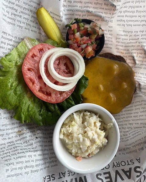 Ein frisch zubereiteter Cheeseburger wird aufgeklappt auf bedrucktem Einschlagpapier mit Zeitungsoptik im berühmten The Salty Dog Cafe in Hilton Head Island, South Carolina, serviert. Auf dem Papier liegt ein saftiges Rinder-Patty mit geschmolzenem Cheddar-Käse neben den separaten Beilagen: einem knackigen grünen Salatblatt, einer großen Tomatenscheibe, frischen Zwiebelringen, einer Gewürzgurke und einem kleinen Schälchen Pico de Gallo sowie einer weißen Keramikschale mit cremigem Coleslaw-Krautsalat im Vordergrund.