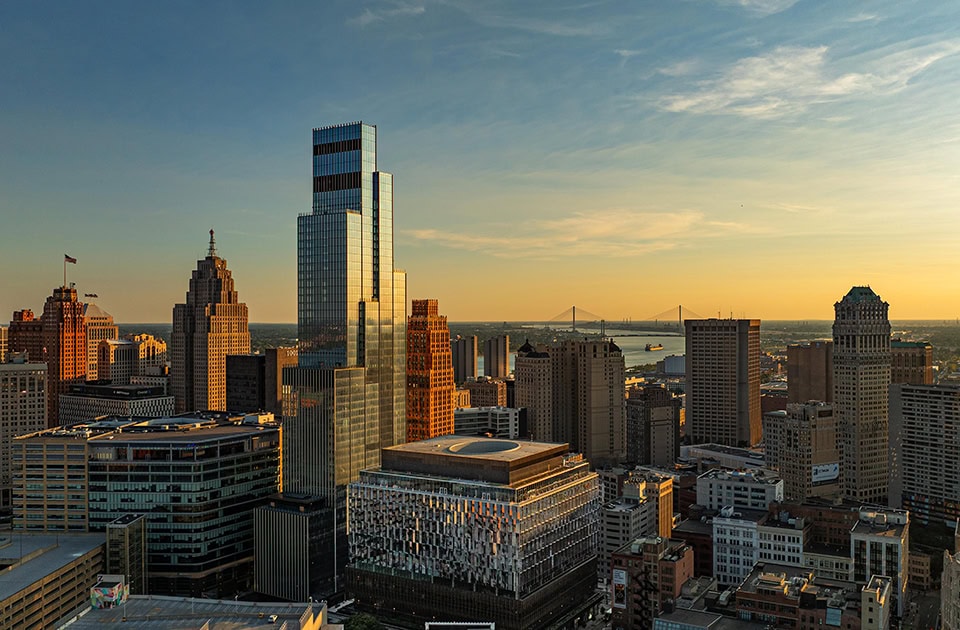 Eine weitläufige Skyline von Detroit im warmen Licht des Sonnenuntergangs, dominiert von modernen Wolkenkratzern und historischen Gebäuden, mit einer Hängebrücke über dem Fluss im fernen Hintergrund.