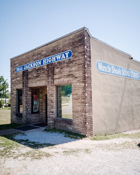 Ein einstöckiges, quaderförmiges Gebäude mit einer gemauerten Sandsteinfront zeigt ein blaues Schild mit der Aufschrift „3614 JACKSON HIGHWAY“ über dem Eingang und ein weiteres Schild an der Seitenwand mit der Aufschrift „Muscle Shoals SOUND STUDIOS“ unter blauem Himmel.