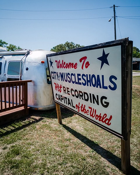 Ein großes Holzschild mit der Aufschrift „Welcome To CITY of MUSCLE SHOALS - HIT RECORDING CAPITAL of the World“ steht auf einer Wiese vor einem silbernen Airstream-Wohnwagen unter blauem Himmel.