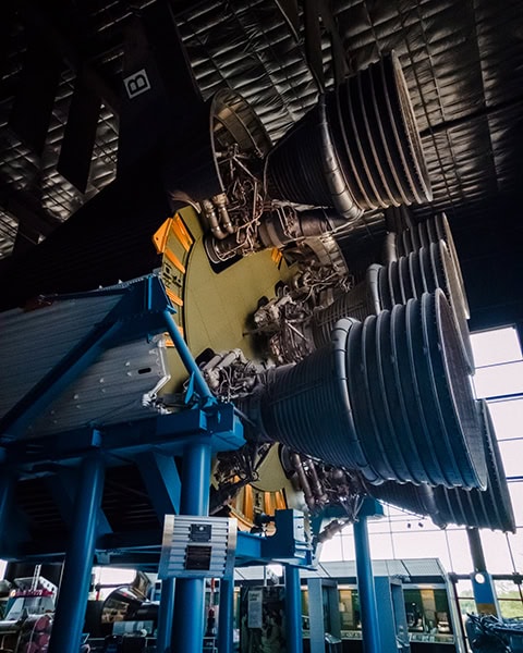 Die gewaltigen, geriffelten Triebwerksdüsen einer historischen Saturn-V-Rakete sind aus einer tiefen Perspektive in einer großen Ausstellungshalle zu sehen, gestützt von einer massiven blauen Metallkonstruktion.