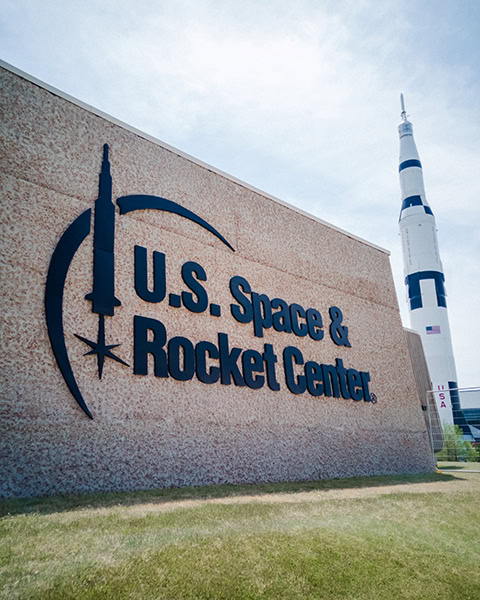 Eine große, strukturierte Betonwand zeigt das Logo und den schwarzen Schriftzug „U.S. Space & Rocket Center“, während im Hintergrund eine riesige, weiß-schwarze Saturn-V-Rakete in den leicht bewölkten Himmel ragt.