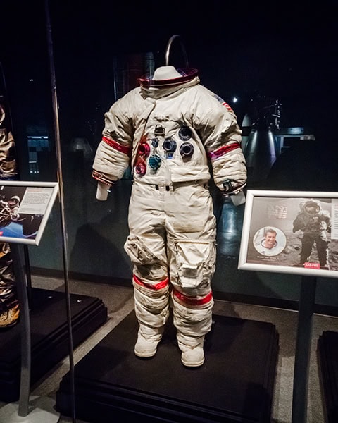 Ein klobiger, weißer Astronautenanzug aus der Apollo-Ära steht ohne Helm auf einem Podest in einer abgedunkelten Museumsausstellung, flankiert von zwei Infotafeln in silbernen Rahmen.
