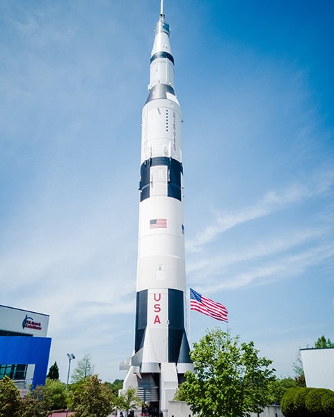 Eine riesige, weiß-schwarze Saturn-V-Rakete ragt senkrecht in den blauen Himmel, während daneben eine US-amerikanische Flagge im Wind weht und im Hintergrund Teile des U.S. Space & Rocket Center Museums zu sehen sind.