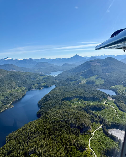 Blick aus dem Fenster eines Flugzeugs, bei dem oben rechts ein Teil des Triebwerks oder der Tragfläche zu sehen ist. Darunter erstreckt sich eine weite Berglandschaft mit tiefblauen, langgestreckten Seen und dichten, grünen Nadelwäldern unter strahlend blauem Himmel.