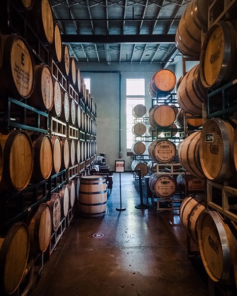 In einer langen, hohen Lagerhalle der St. Augustine Distillery in St. Augustine, Florida, stapeln sich zahlreiche traditionelle Bourbon- und Whiskey-Holzfässer auf mehrstöckigen Metallregalen entlang der Wände. Ein zentraler Gang führt vorbei an den Holzfässern hin zu zwei hellen Fenstern im Hintergrund, während ein einzelnes Ausstellungsfass und Absperrständer den Besucherbereich markieren.