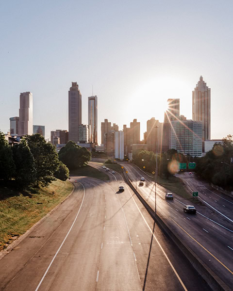 Die Skyline von Downtown Atlanta erstreckt sich hinter einer mehrspurigen Autobahn, auf der sich einige Autos bewegen, während die tief stehende Sonne hinter einem der Wolkenkratzer hervorstrahlt und lange Schatten wirft.
