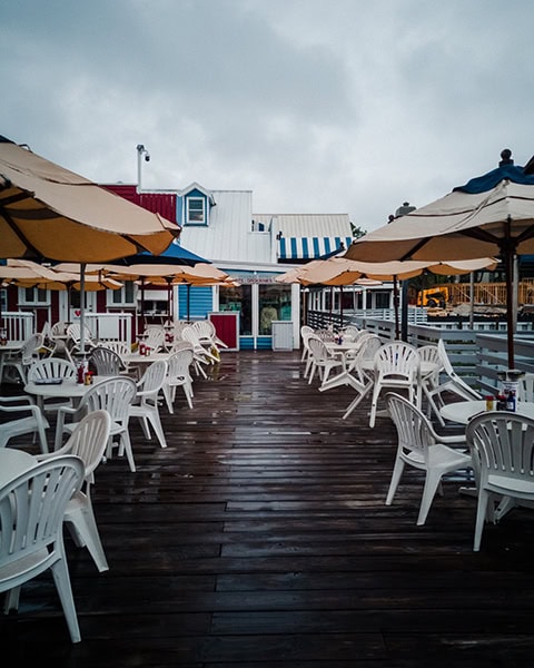 Die nasse, hölzerne Außenterrasse des berühmten The Salty Dog Cafe in der South Beach Marina auf Hilton Head Island, South Carolina, ist mit zahlreichen weißen Plastiktischen und -stühlen unter aufgespannten, beigen Sonnenschirmen möbliert, während im Hintergrund die charakteristischen bunten und blau-weiß gestreiften Gebäude des Hafenviertels unter einem bewölkten Himmel zu sehen sind.