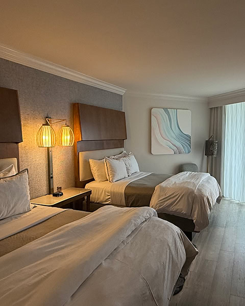 Ein modern und elegant eingerichtetes Hotelzimmer im The Westin Hilton Head Island Resort & Spa zeigt zwei große, gemütlich hergerichtete Doppelbetten mit weißen Decken und Kissen vor hohen Holzkopfteilen an einer grauen Strukturtatpete, beleuchtet von einer warm strahlenden Nachttischlampe im Korbdesign, während im Hintergrund ein abstraktes Wellengemälde an der Wand hängt und Tageslicht durch ein großes Fenster mit transparenten Vorhängen auf den hellen Holzboden fällt.
