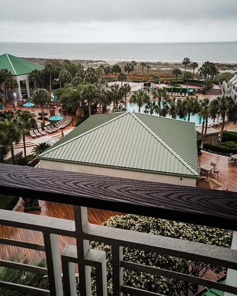 Ein Blick von einem Hotelbalkon mit gemustertem weißen Geländer zeigt die weitläufige Pool- und Gartenlandschaft des luxuriösen The Westin Hilton Head Island Resort & Spa auf Hilton Head Island, South Carolina, mit Palmen, Liegestühlen, einem großen Poolbereich und einem Gebäude mit grünem Walmdach im Vordergrund, während sich im Hintergrund der Sandstrand und das Meer unter einem bewölkten Himmel erstrecken.