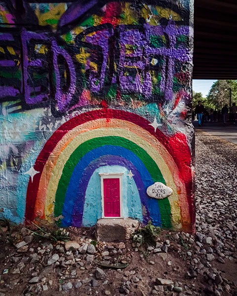 Eine kleine, pinkfarbene Miniaturtür ist am Fuß einer mit Graffiti besprühten Betonwand eingelassen, umrahmt von einem bunten, aufgemalten Regenbogen und einem kleinen Schild mit der Aufschrift „Tiny Doors ATL“.