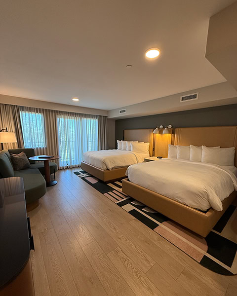 Ein geräumiges, modernes Hotelzimmer mit hellem Holzboden und zwei großen Doppelbetten mit weißer Bettwäsche. Links steht ein geschwungenes, dunkelgrünes Sofa mit einem kleinen Holztisch, im Hintergrund hängen bodenlange, beige Vorhänge vor großen Fenstern.
