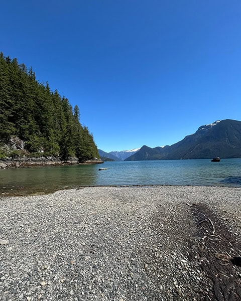 Ein weiter Kieselstrand im Vordergrund führt zu einem ruhigen, blauen See. Links ragt eine steile, dicht mit grünen Nadelbäumen bewachsene Küstenlinie empor. Im Hintergrund erstrecken sich majestätische Berge mit kleinen Schneefeldern unter einem wolkenlosen, tiefblauen Himmel.