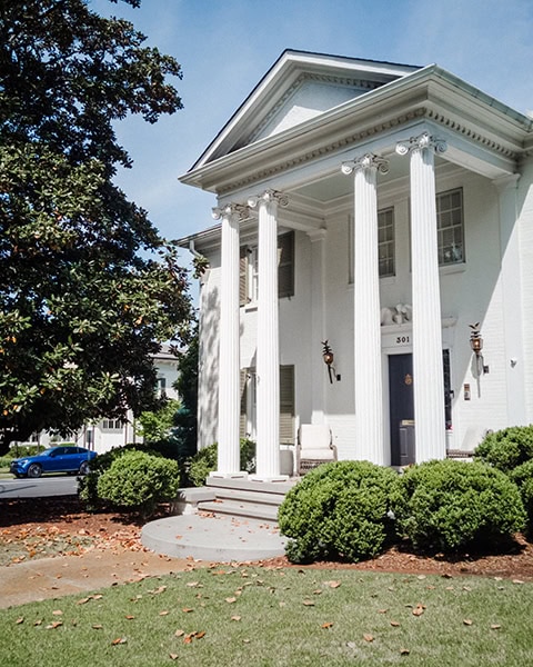 Eine große, weiße Villa im neoklassizistischen Antebellum-Stil mit markanten ionischen Säulen und einem dreieckigen Giebel steht hinter einem gepflegten Vorgarten mit grünen Büschen unter blauem Himmel.