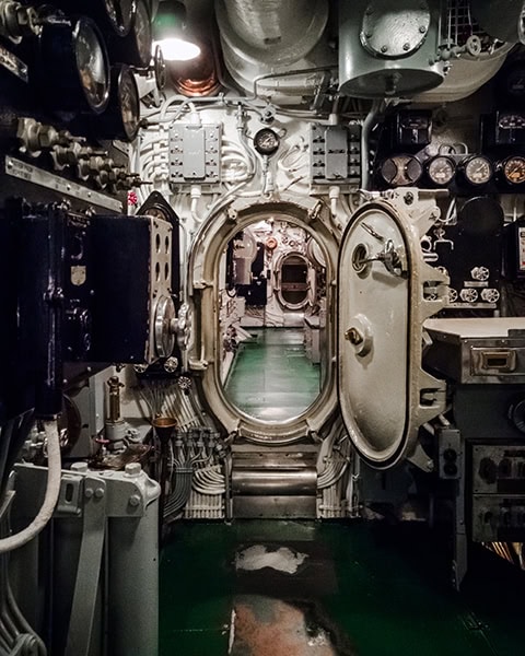 Ein Blick durch ein geöffnetes, massives Rundschott führt in die engen Gänge des historischen U-Boots USS Drum (SS-228) im USS Alabama Battleship Memorial Park in Mobile, umgeben von zahlreichen Leitungen, Ventilen, Schaltern und Druckanzeigen an den Wänden.