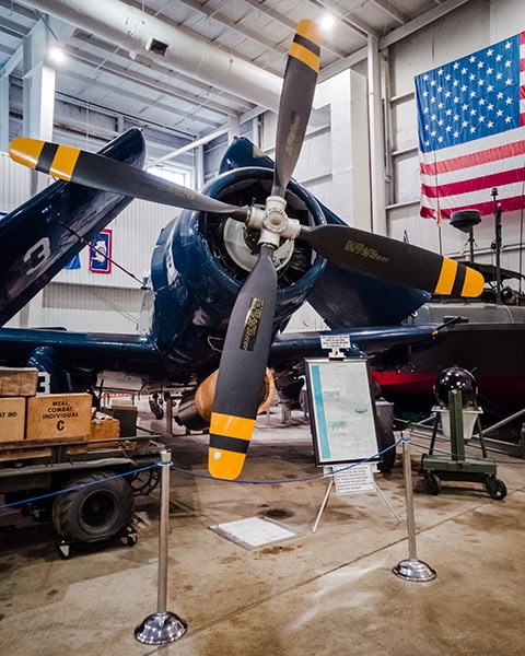 Ein historisches Kampfflugzeug vom Typ Vought F4U Corsair mit hochgeklappten Tragflächen steht im USS Alabama Battleship Memorial Park (im dortigen Pavilion für Militärflugzeuge) in Mobile, während im Hintergrund eine große US-amerikanische Flagge von der Decke hängt und im Vordergrund eine Infotafel sowie Absperrpfosten zu sehen sind.