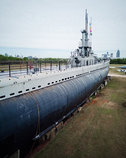 Das historische U-Boot USS Drum (SS-228) aus dem Zweiten Weltkrieg liegt aufgebockt auf einer Rasenfläche im USS Alabama Battleship Memorial Park in Mobile, mit Blick auf das lange Oberdeck, den Kommandoturm mit seinen Antennen und die Skyline von Mobile in der Ferne unter bewölktem Himmel.