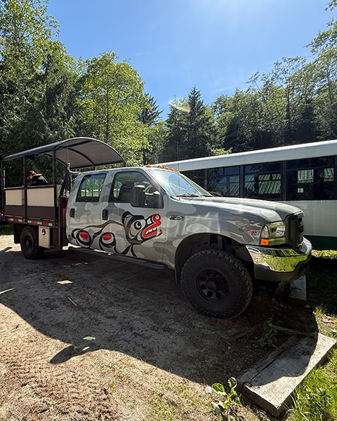 Ein großer, hellgrauer Pickup-Truck mit Camouflage-Muster und einer Doppelkabine steht im Sonnenschein auf unbefestigtem Boden vor dichten grünen Bäumen. Auf der Seite des Trucks ist eine traditionelle Bärengrafik im rot-schwarzen First-Nations-Stil aufgemalt.