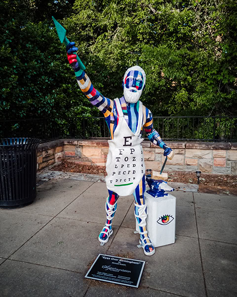 Eine bunt bemalt Statue einer bärtigen, männlichen Figur im Stil des Gottes Vulcan trägt eine weiße Schürze, die als Sehtest-Tafel mit Buchstaben gestaltet ist, und hält einen grünen Speilaufwärts gestreckt, während sie neben einem Amboss im Freien steht.