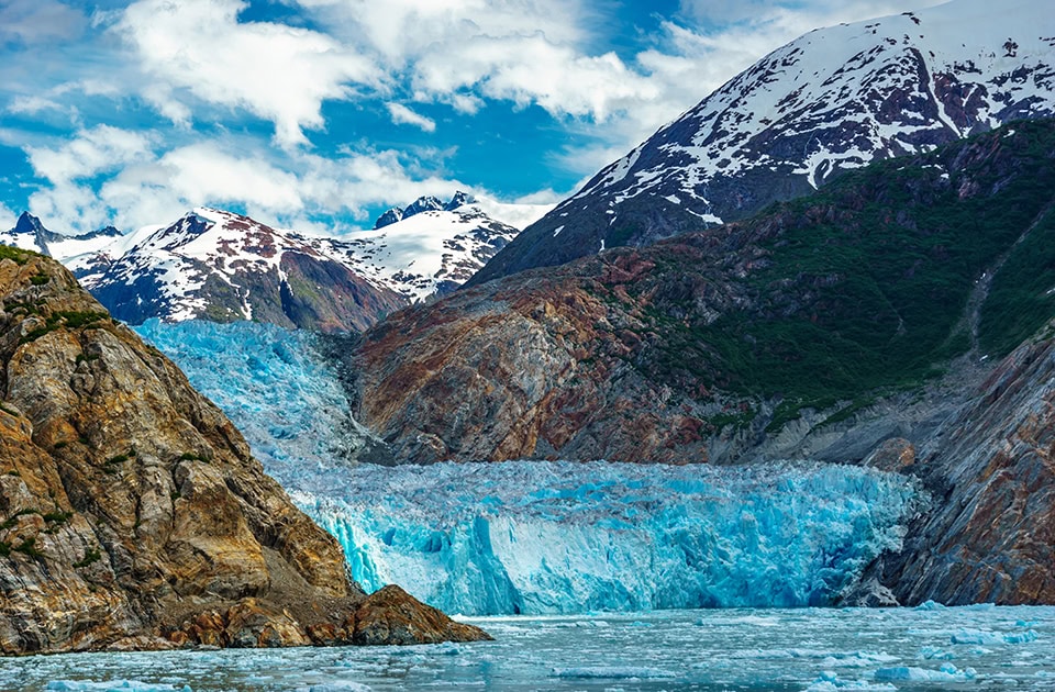 Eine Nahaufnahme des leuchtend blauen Sawyer-Gletschers im Tracy Arm Fjord in Alaska. Das zerklüftete, intensiv hellblaue Gletschereis mündet direkt in ein mit kleinen Eisbergen und Eisschollen bedecktes Gewässer im Vordergrund. Der Gletscher ist von steilen, felsigen und teils braun-grauen Berghängen umgeben. Im Hintergrund ragen hohe, schneebedeckte Berggipfel unter einem teilweise bewölkten blauen Himmel empor.