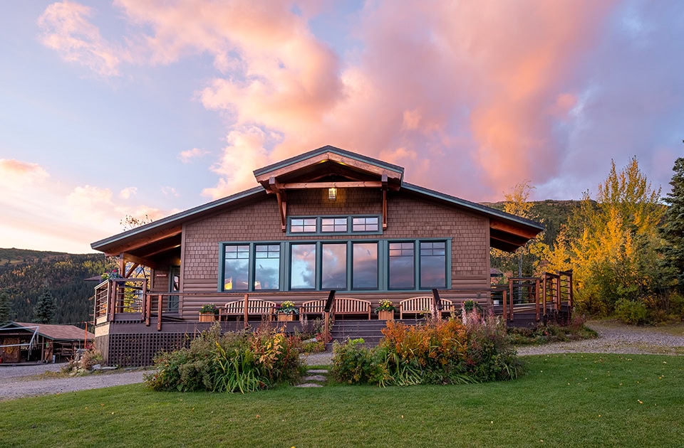 Eine einstöckige Lodge aus Holz mit einer Terrasse bei Sonnenuntergang unter einem rosa-orange leuchtenden Wolkenhimmel.