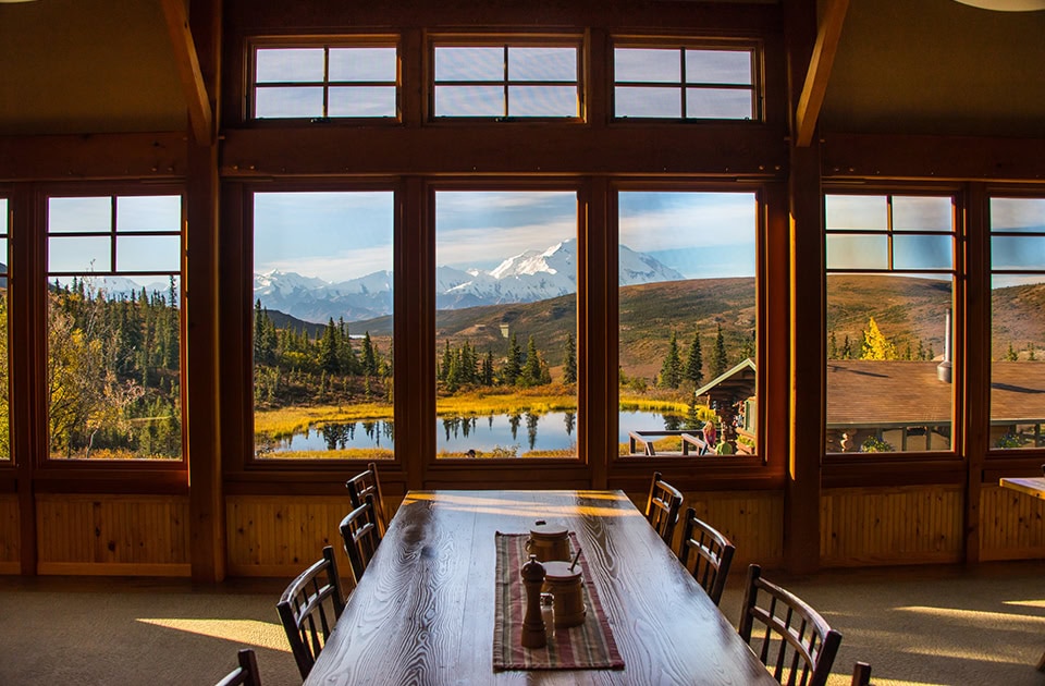 Innenansicht einer Lodge mit Blick durch eine große Fensterfront auf einen See und schneebedeckte Berge.