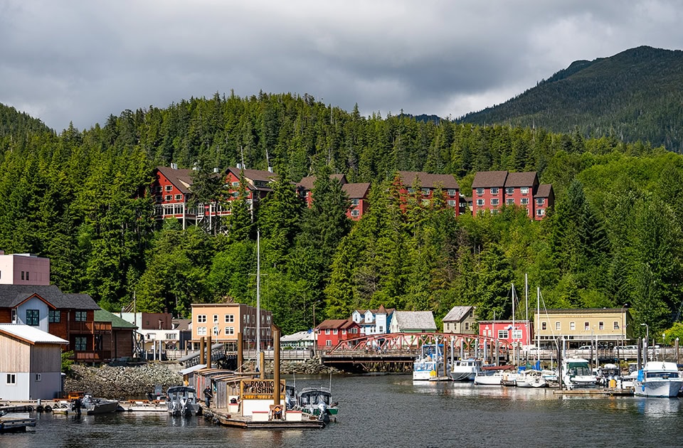 Blick auf den Hafen Thomas Basin in Ketchikan, Alaska, mit zahlreichen Booten an den Stegen im Vordergrund. Dahinter erstreckt sich die Stadt mit einer markanten roten Brücke. Ein dichter, grüner Nadelwald bedeckt den Hügel im Hintergrund, auf dem ein großer, roter Hotelkomplex (die Cape Fox Lodge) thront. Der Himmel ist bewölkt.
