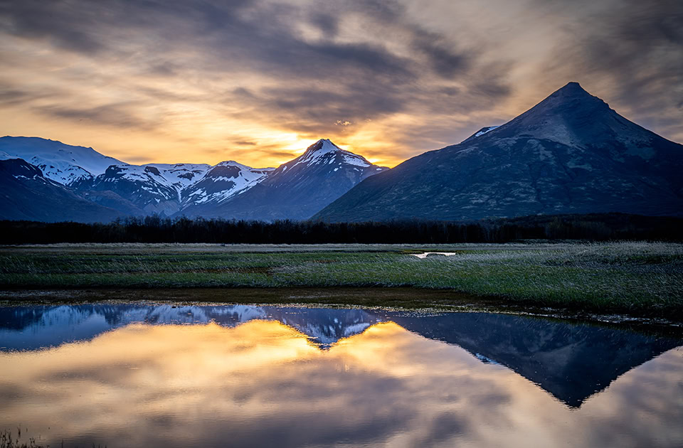 Eine weite Landschaft bei Sonnenuntergang mit Bergen, die sich in einer ruhigen Wasserfläche im Vordergrund spiegeln. Rechts ragt ein markanter, spitzer Berg empor, während links schneebedeckte Berggipfel zu sehen sind. Der Himmel ist dramatisch bewölkt und hinter den Bergen leuchtet ein goldener Sonnenuntergang.