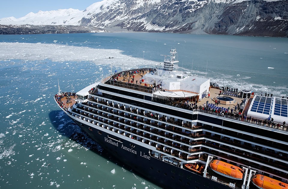Ein großes Kreuzfahrtschiff der „Holland America Line“ fährt durch ein eisiges Gewässer mit zahlreichen kleinen Eisschollen, während im Hintergrund schneebedeckte Berge aufragen.