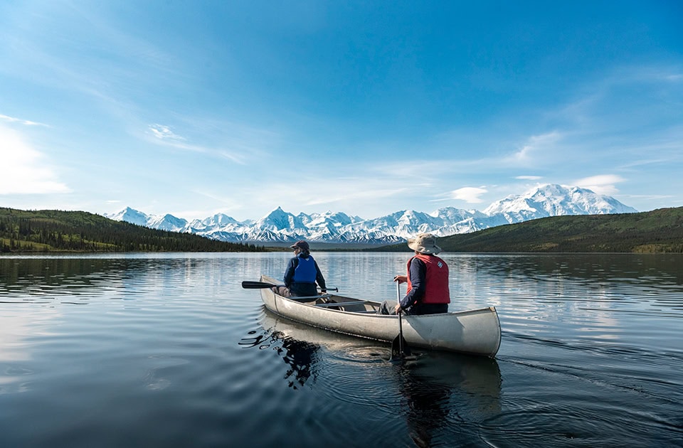 Zwei Personen paddeln in einem Kanu auf einem ruhigen See vor einer schneebedeckten Bergkette unter blauem Himmel.