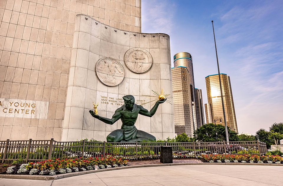 Die große, grüne Bronzestatue „The Spirit of Detroit“ sitzt vor einer hellen Marmorwand des Coleman A. Young Municipal Center. Sie hält eine goldene Figurengruppe in der rechten und eine goldene Kugel mit Strahlen in der linken Hand. Im Hintergrund ragen die modernen Wolkenkratzer des Renaissance Center unter einem leicht bewölkten, blauen Himmel empor.