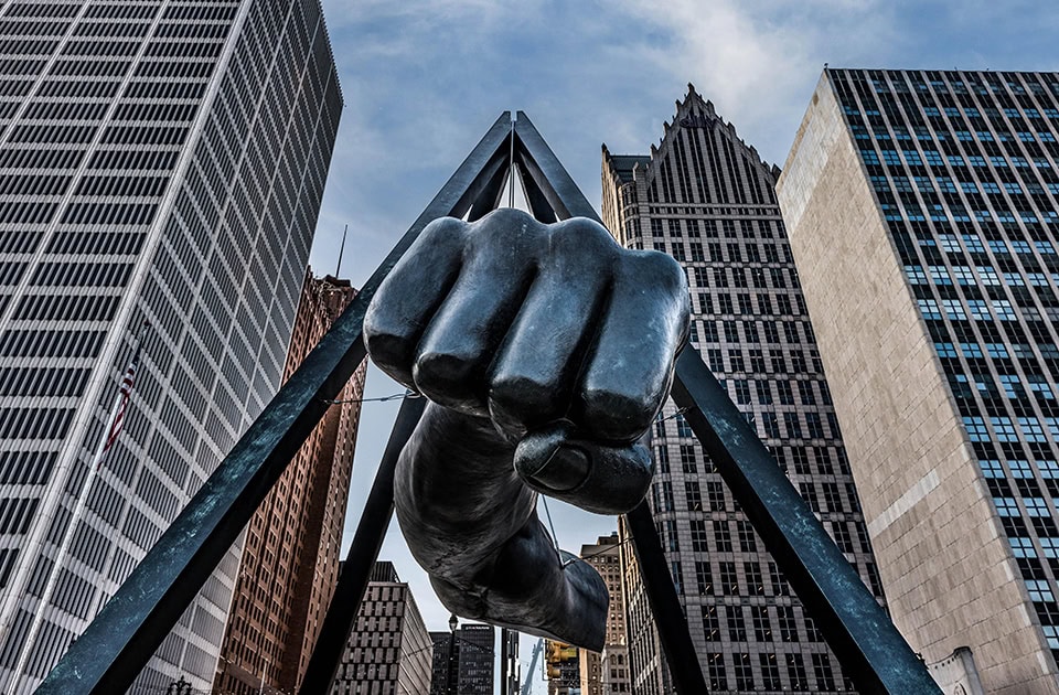 Eine monumentale Skulptur einer geballten, schwarzen Faust, die an einem pyramidenartigen Metallgerüst aufgehängt ist und vor einer Kulisse aus modernen Wolkenkratzern und blauem Himmel aufragt.