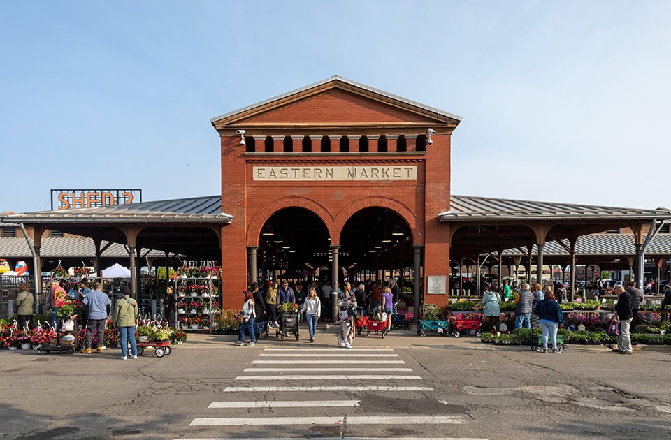 Ein markantes, zentrales Backsteingebäude mit zwei großen Rundbögen und der Aufschrift „EASTERN MARKET“ an einem sonnigen Tag. Links schließt sich eine offene Markthalle mit dem Schild „SHED 2“ an. Zahlreiche Menschen besuchen den belebten Markt, auf dem viele bunte Blumen und Pflanzen im Freien ausgestellt sind. Im Vordergrund befindet sich ein Zebrastreifen auf dem Asphalt.