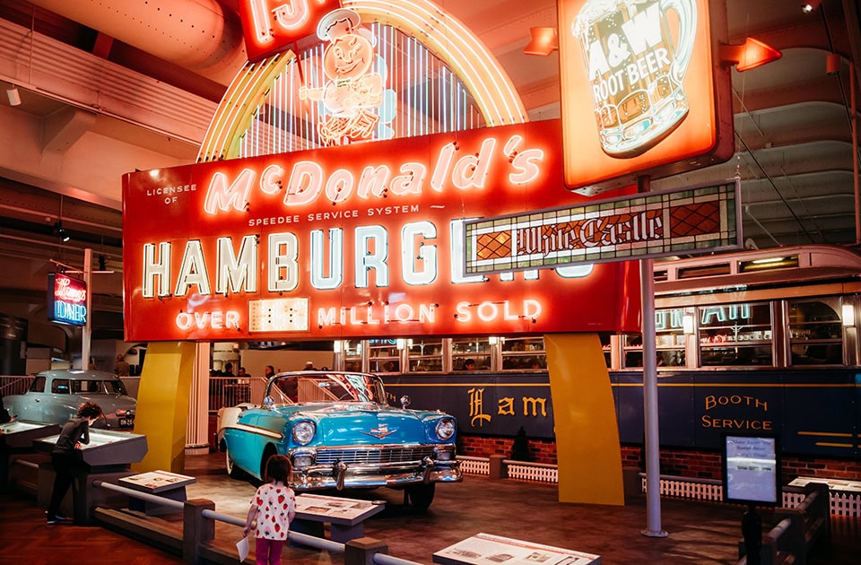 Ein historisches, leuchtend rotes McDonald’s-Neonschild mit der Aufschrift „McDonald’s Hamburgers - Over 1 Million Sold“ dominiert die Szene in einer Museumshalle. Vor dem Schild steht ein Oldtimer-Cabriolet in Hellblau und Weiß. Im Hintergrund sind weitere Nostalgie-Exponate wie ein „A&W Root Beer“-Schild und ein historischer Diner-Wagen zu sehen, während Museumsbesucher die Ausstellung betrachten.