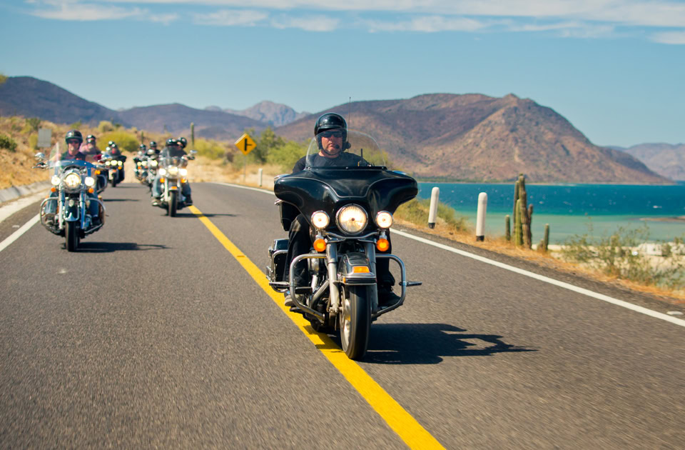 Eine Gruppe von Bikern fährt auf Harley-Davidson-Motorrädern über eine asphaltierte Landstraße in Baja California Sur, Mexiko. Der führende Fahrer steuert eine schwarze Touring-Maschine mit großer Verkleidung direkt auf der gelben Mittellinie. Im Hintergrund erstrecken sich das türkisfarbene Wasser der Conception Bay und karge, rötlich-braune Wüstenberge unter einem leicht bewölkten Himmel.