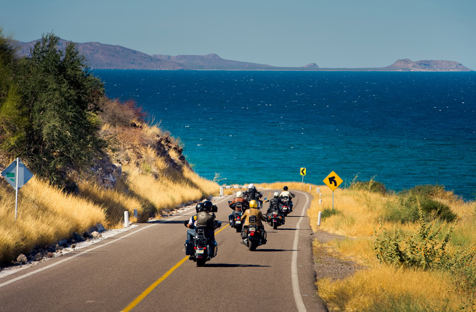 Eine Gruppe von Motorradfahrern fährt hintereinander eine asphaltierte Straße hinab, die geradewegs auf das tiefblaue Meer der Conception Bay (Bahía de Concepción) in Baja California Sur, Mexiko, zuführt. Am rechten Straßenrand stehen gelbe Verkehrsschilder, die vor einer scharfen Linkskurve warnen, während im Hintergrund eine karge Hügelkette am Horizont aufragt.