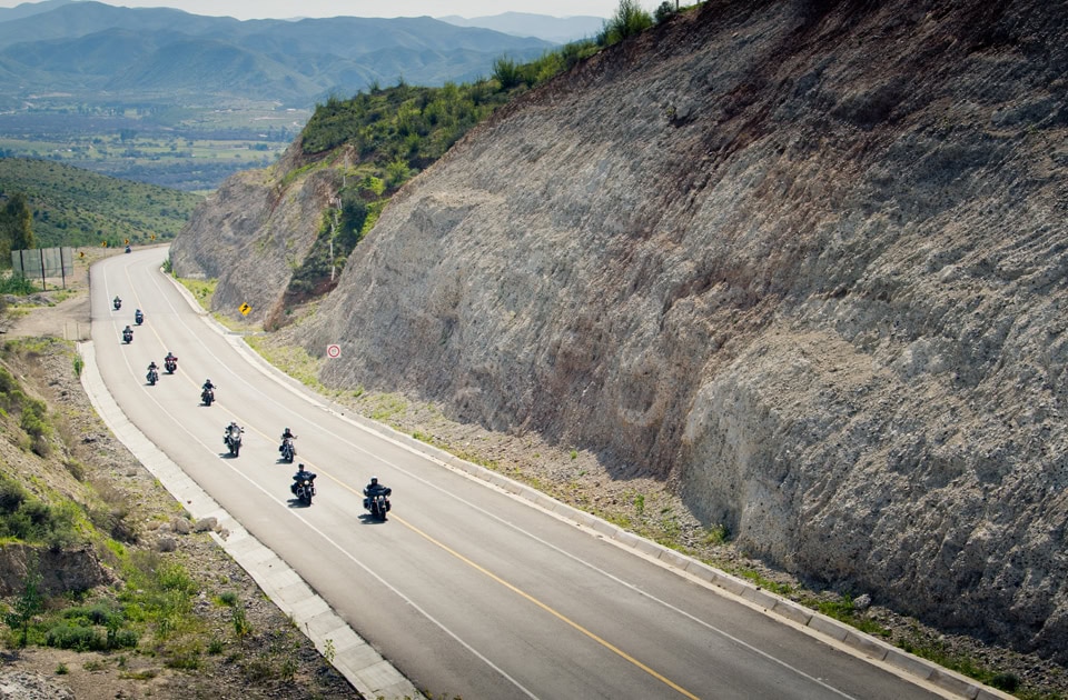 Eine Kolonne von Motorradfahrern fährt hintereinander eine breite, kurvige Bergstraße hinab, die sich durch die karge Gebirgslandschaft der Baja California in Mexiko zieht. Die Straße verläuft entlang einer steilen, hellen Felswand auf der rechten Seite, während sich im Hintergrund weite Täler und sanfte Hügelketten unter einem leicht dunstigen Himmel erstrecken.