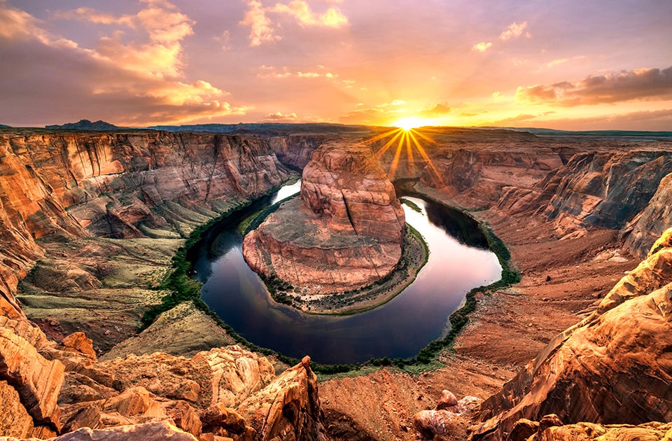 Der Colorado River windet sich in einer hufeisenförmigen Kurve, der sogenannten Horseshoe Bend, um eine gewaltige, rotbraune Felsformation in einem tiefen Canyon. Am Horizont geht die Sonne unter und wirft strahlenförmiges, goldenes Licht über die gesamte Schlucht und den leicht bewölkten Himmel.