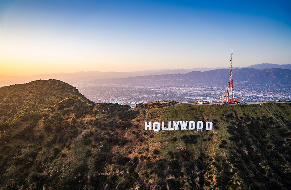 Der weltberühmte weiße „HOLLYWOOD“-Schriftzug erstreckt sich auf der grünen Flanke des Mount Lee. Rechts daneben steht ein hoher, rot-weißer Sendemast auf dem Berggipfel, während im Hintergrund das weitläufige Stadtgebiet von Los Angeles und dunstige Bergketten im sanften Licht der Abenddämmerung zu sehen sind.
