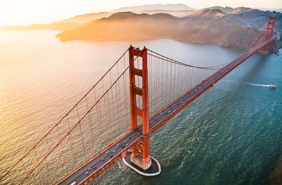 Die rot-orangefarbene Golden Gate Bridge erstreckt sich aus einer hohen Perspektive über das tiefblaue Wasser der Meeresstraße. Ein Brückenpfeiler steht markant im Vordergrund, während die hügelige Küstenlandschaft der Marin Headlands im Hintergrund vom goldenen Licht der tiefstehenden Sonne angestrahlt wird.