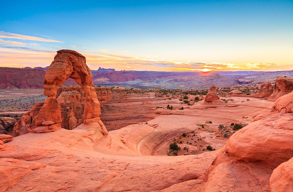 Der freistehende, rotbraune Felsbogen des Delicate Arch ragt auf einer glatten Sandsteinfläche über einer weiten Wüstenschlucht empor. Im Hintergrund geht die Sonne am Horizont unter und färbt den Himmel in sanften Orange-, Gelb- und Blautönen.