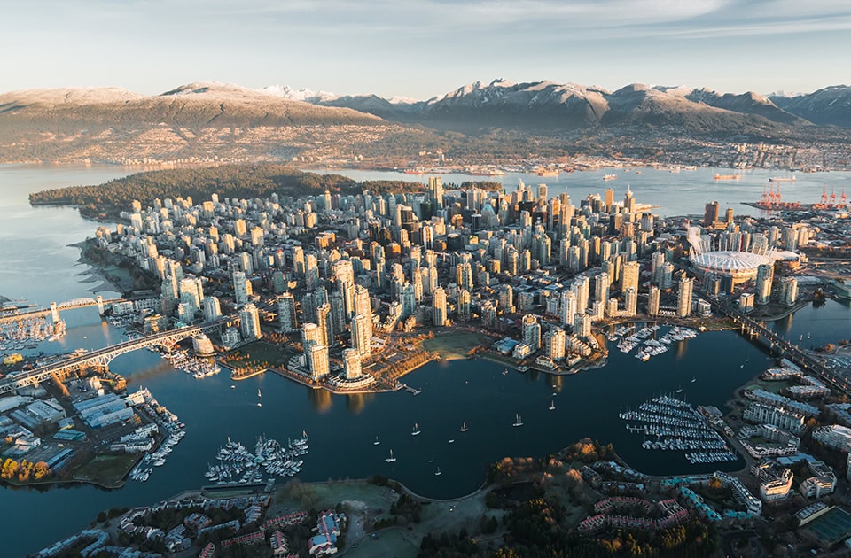 Eine Luftaufnahme der Skyline von Vancouver, Kanada, bei Sonnenaufgang oder Sonnenuntergang. Die dicht bebaute Halbinsel Downtown Vancouver ist von Wasser umgeben. Im Hintergrund ragen die schneebedeckten North Shore Mountains unter einem klaren Himmel empor. Das goldene Licht der tiefstehenden Sonne spiegelt sich auf den Glasfassaden der Wolkenkratzer und den angrenzenden Buchten False Creek und Burrard Inlet.