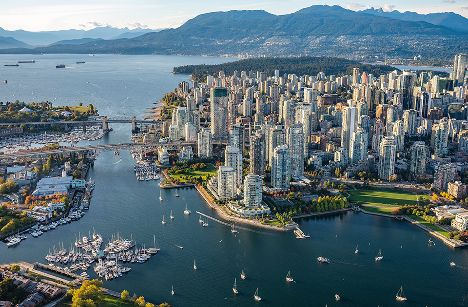 Luftaufnahme der Skyline von Vancouver mit zahlreichen Wolkenkratzern, die an eine von Segelbooten und Brücken umgebene Bucht grenzen, im Hintergrund bewaldete Berge unter blauem Himmel.