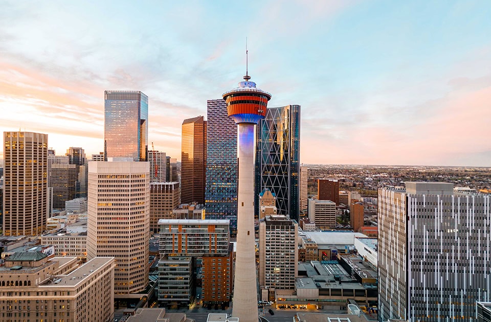 Der markante Calgary Tower mit seiner rot-weißen Aussichtsplattform steht im Zentrum einer modernen Skyline mit zahlreichen Wolkenkratzern während eines farbenfrohen Sonnenuntergangs mit rosa-orangefarbenen Wolken.