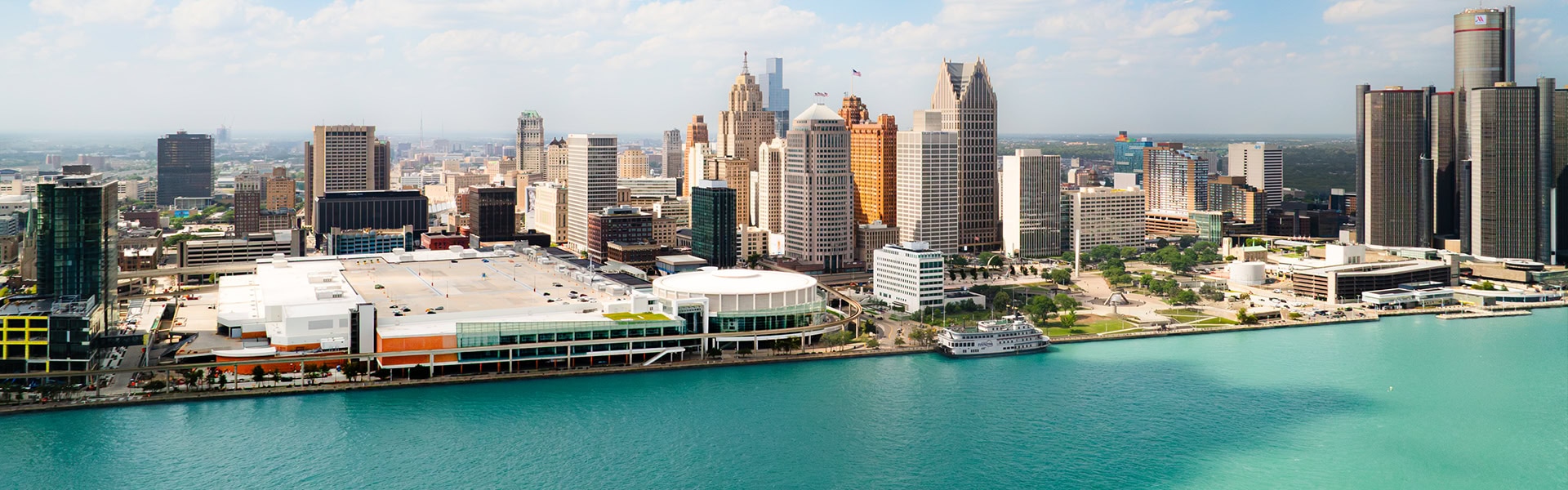 Ein Panorama der Skyline von Detroit, Michigan, aufgenommen aus der Vogelperspektive über dem Detroit River.Im Vordergrund erstreckt sich das tiefblaue bis türkisfarbene Wasser des Flusses. Am Ufer zieht sich die Uferpromenade entlang, an der markante Gebäude wie das weitläufige Huntington Place Kongresszentrum (mit dem runden Atrium) und der Plaza-Bereich zu sehen sind. Dahinter reihen sich die Wolkenkratzer der Innenstadt aneinander, darunter das markante, spitze One Detroit Center (Ally Detroit Center) und ganz rechts der monumentale Komplex des Renaissance Center (GM-Hauptquartier). Über der Stadt erstreckt sich ein strahlend blauer Himmel mit vielen weißen Schäfchenwolken.
